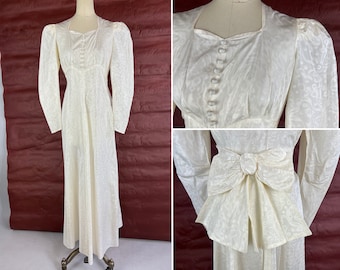 May include: A white vintage wedding dress with a fitted bodice, long sleeves, and a full skirt. The dress features a square neckline, a button-up front, and a large bow at the back.