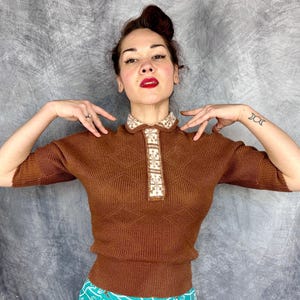 Pu&ograve; includere: Un top in maglia marrone con scollo a camicia e maniche corte. Il colletto e la paramontura presentano un motivo geometrico color crema e beige. Il top ha un motivo a rombi ed &egrave; indossato da una persona.