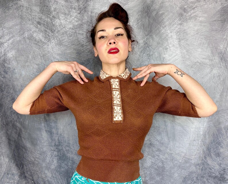 Pu&ograve; includere: Un top in maglia marrone con scollo a camicia e maniche corte. Il colletto e la paramontura presentano un motivo geometrico color crema e beige. Il top ha un motivo a rombi ed &egrave; indossato da una persona.