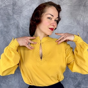 May include: Mustard yellow blouse with a keyhole neckline and long, gathered sleeves. The blouse features a mandarin collar and decorative stitching around the keyhole. The sleeves have elastic cuffs. The blouse is made of a lightweight fabric.
