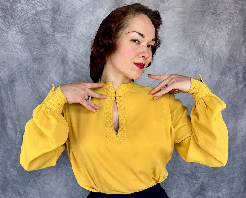 May include: Mustard yellow blouse with a keyhole neckline and long, gathered sleeves. The blouse features a mandarin collar and decorative stitching around the keyhole. The sleeves have elastic cuffs. The blouse is made of a lightweight fabric.