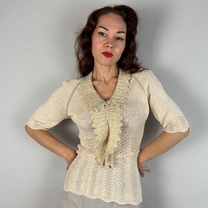 May include: A cream-colored crocheted sweater with a delicate lace trim and a ruffled neckline. The sweater has a fitted silhouette and a short sleeve.