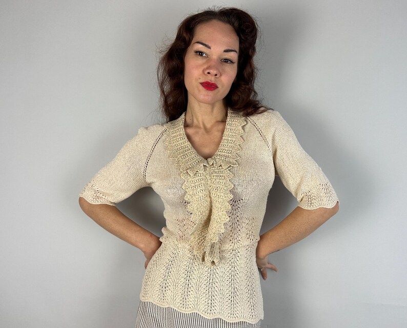 May include: A cream-colored crocheted sweater with a delicate lace trim and a ruffled neckline. The sweater has a fitted silhouette and a short sleeve.