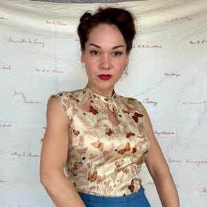 May include: A sleeveless, cream-colored blouse with a keyhole neckline. The blouse features a repeating pattern of brown butterflies and floral elements. The model is wearing blue pants. The background is a cream-colored fabric with handwritten names.