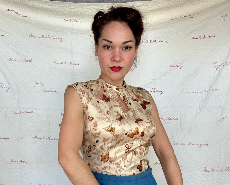 May include: A sleeveless, cream-colored blouse with a keyhole neckline. The blouse features a repeating pattern of brown butterflies and floral elements. The model is wearing blue pants. The background is a cream-colored fabric with handwritten names.