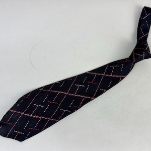 Puede incluir: Una corbata azul marino con un patrón repetitivo de guiones y líneas rojas y blancas. La corbata está anudada cerca de la parte superior y se encuentra en diagonal sobre una superficie blanca.