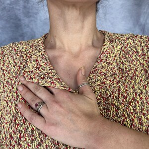 May include: A textured knit jacket in yellow, red, and brown. The jacket has a collared neckline and is buttoned. A hand with rings is placed over the jacket.