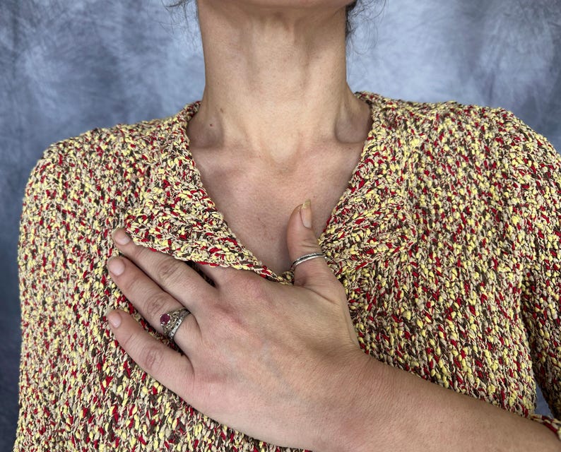 May include: A textured knit jacket in yellow, red, and brown. The jacket has a collared neckline and is buttoned. A hand with rings is placed over the jacket.