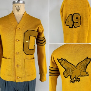 Könnte beinhalten: Ein Vintage-Gelb-Cardigan mit einem großen Filzbuchstaben "C" auf dem linken Ärmel und einem Filzadler auf der Rückseite. Der Cardigan hat zwei Taschen und einen Knopfleisten vorne. Die Ärmel haben schwarze Streifen.