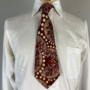 May include: A patterned tie and white shirt. The tie has a design of maroon and tan radial shapes and white polka dots. The white shirt has a button-down collar and a pocket on the left side.