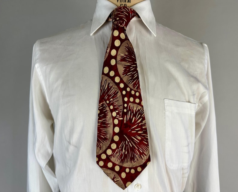 May include: A patterned tie and white shirt. The tie has a design of maroon and tan radial shapes and white polka dots. The white shirt has a button-down collar and a pocket on the left side.