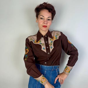May include: A long-sleeve button-down shirt in dark brown with tan panels on the shoulders and cuffs. The shirt features colourful floral embroidery in yellow, orange, and blue on the chest, shoulders, and cuffs, with a pointed collar and buttons.