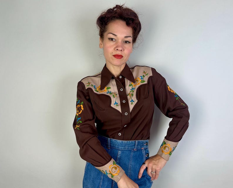 May include: A long-sleeve button-down shirt in dark brown with tan panels on the shoulders and cuffs. The shirt features colourful floral embroidery in yellow, orange, and blue on the chest, shoulders, and cuffs, with a pointed collar and buttons.