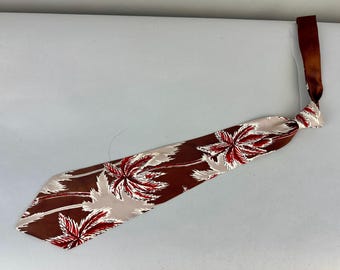 May include: A necktie featuring a dark brown background with a repeating pattern of stylized palm tree leaves in white and deep red. The tie has a pointed end and a matching dark brown neck strap with a knot detail at the top of the main blade.