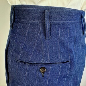 Puede incluir: Pantalones azules de talle alto con un sutil patr&oacute;n de rayas diplom&aacute;ticas blancas. La prenda incluye una cintura estructurada con trabillas para cintur&oacute;n y un bolsillo trasero ribeteado con un bot&oacute;n oscuro de superficie texturizada.