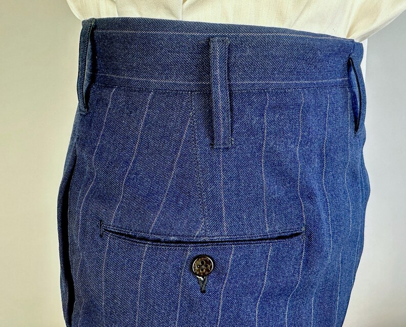 Puede incluir: Pantalones azules de talle alto con un sutil patr&oacute;n de rayas diplom&aacute;ticas blancas. La prenda incluye una cintura estructurada con trabillas para cintur&oacute;n y un bolsillo trasero ribeteado con un bot&oacute;n oscuro de superficie texturizada.