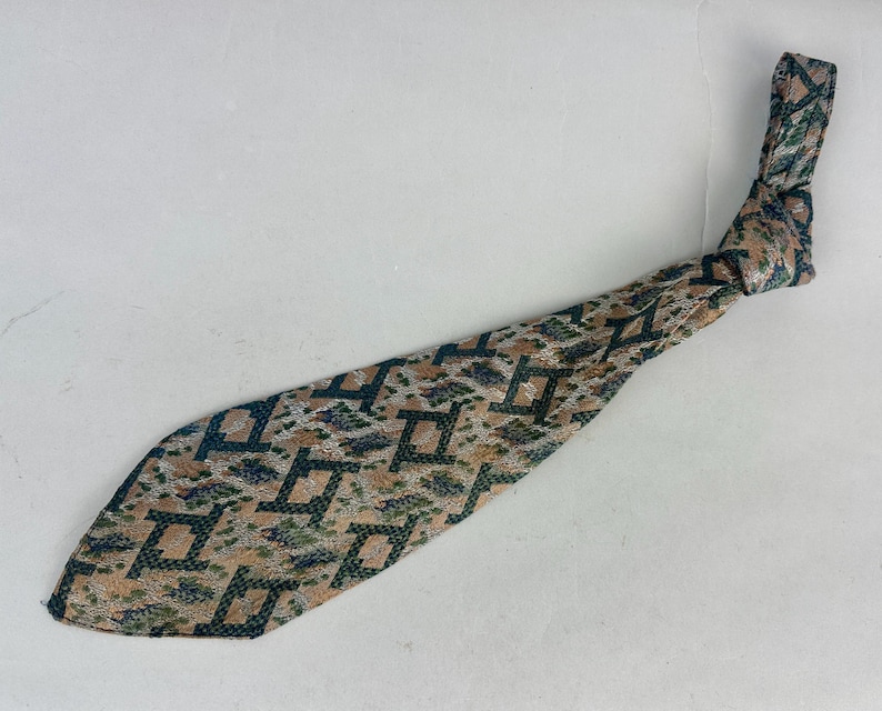 May include: A necktie featuring a repeating geometric pattern of dark blue squares on a tan background, accented with small patches of green and silver textured fabric. The tie is pre-knotted and displays a woven finish throughout the entire surface.