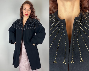 May include: A black open-front coat with gold-colored embellishments on the shoulders and lapels. The coat has long sleeves and pockets. The coat is worn over a pink dress.