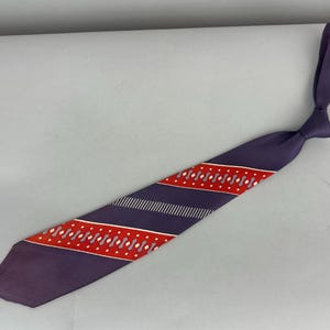 Può includere: Una cravatta viola con un motivo geometrico rosso e bianco.