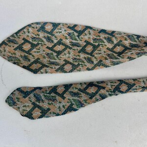 May include: A necktie featuring a repeating geometric pattern in dark green, tan, and silver. The fabric has a textured appearance with sharp, pointed ends. The design consists of interconnected square shapes layered over a mottled background.