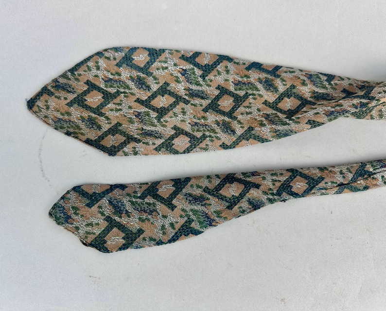 May include: A necktie featuring a repeating geometric pattern in dark green, tan, and silver. The fabric has a textured appearance with sharp, pointed ends. The design consists of interconnected square shapes layered over a mottled background.