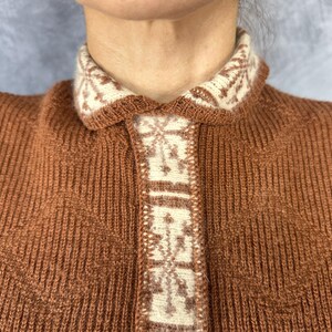 Pu&ograve; includere: Primo piano di un cardigan marrone a maglia con colletto e pannello frontale color crema. Il colletto e il pannello presentano un motivo geometrico color crema e marrone. Il cardigan &egrave; realizzato in tessuto a maglia a coste.