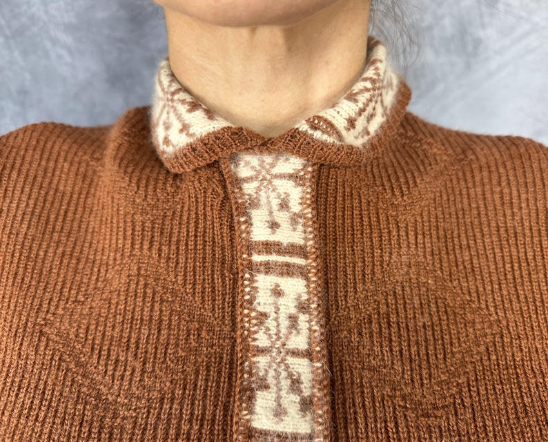Pu&ograve; includere: Primo piano di un cardigan marrone a maglia con colletto e pannello frontale color crema. Il colletto e il pannello presentano un motivo geometrico color crema e marrone. Il cardigan &egrave; realizzato in tessuto a maglia a coste.