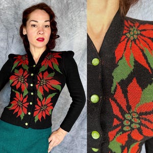 May include: Black cardigan sweater with a poinsettia floral design in red and green. The long-sleeved sweater has a V-neck, shoulder pads, and gold-colored buttons. The woman is wearing a teal green skirt.