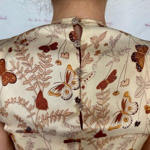 May include: Cream-colored fabric with a butterfly and botanical print. The fabric is buttoned up the back with small, round buttons. The butterflies are brown and orange, and the botanical elements are in shades of brown and tan.
