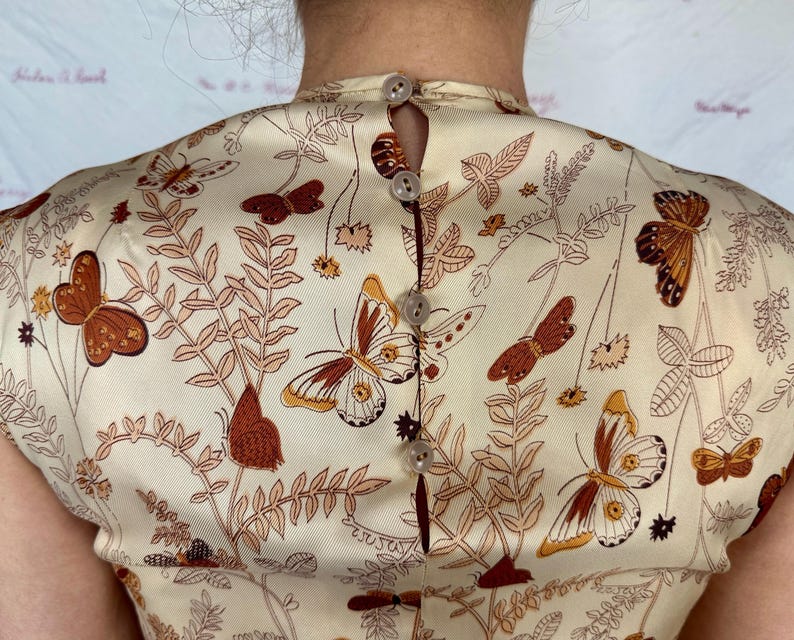 May include: Cream-colored fabric with a butterfly and botanical print. The fabric is buttoned up the back with small, round buttons. The butterflies are brown and orange, and the botanical elements are in shades of brown and tan.
