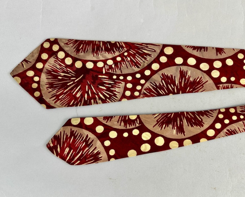 May include: Two neckties with a repeating pattern of red and beige fan-like shapes and white polka dots. The ties are on a white background. The ties are likely made of silk or a similar material.