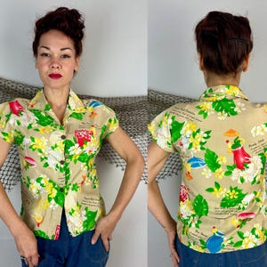 May include: Tan short-sleeved button-up shirt with a tropical floral print featuring red, yellow, and green elements. The shirt has a classic collar and wooden buttons. The print includes illustrations of drinks and text.