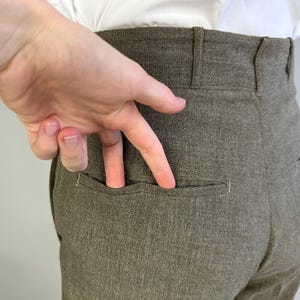 Op de afbeelding: Broek met hoge taille in een effen taupe-bruine kleur, voorzien van een gestructureerde tailleband met riemlussen en een enkele horizontale paspelzak aan de achterkant. De stof heeft een subtiele, fijnkorrelige textuur over het kledingstuk.