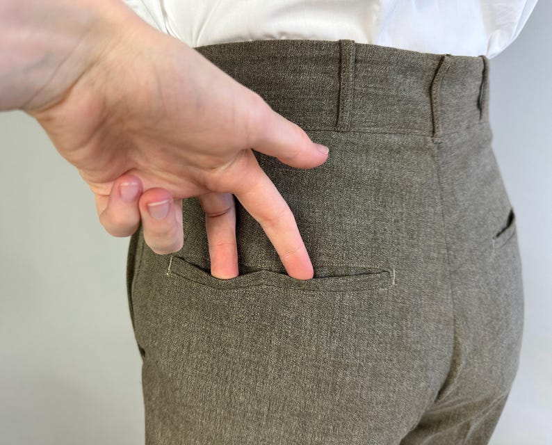 Op de afbeelding: Broek met hoge taille in een effen taupe-bruine kleur, voorzien van een gestructureerde tailleband met riemlussen en een enkele horizontale paspelzak aan de achterkant. De stof heeft een subtiele, fijnkorrelige textuur over het kledingstuk.
