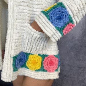 May include: A white textured jacket featuring a horizontal band at the hem and cuffs decorated with embroidered floral motifs in shades of blue, yellow, and pink on a green background. The fabric has a ribbed, plush appearance throughout the garment.