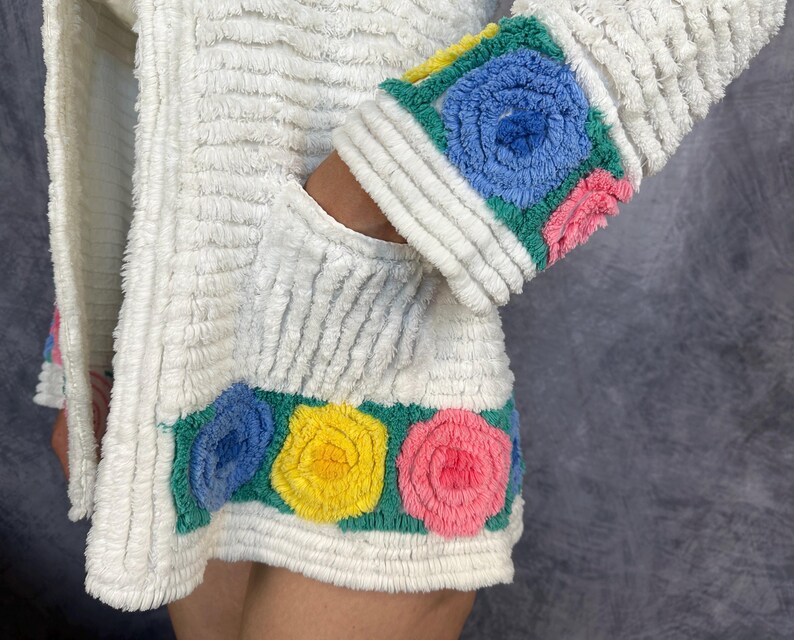 May include: A white textured jacket featuring a horizontal band at the hem and cuffs decorated with embroidered floral motifs in shades of blue, yellow, and pink on a green background. The fabric has a ribbed, plush appearance throughout the garment.
