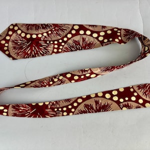 May include: A maroon necktie featuring a repeating pattern of beige half-circles, white polka dots, and maroon spiky shapes. The tie is made of a smooth fabric and is displayed on a white surface.