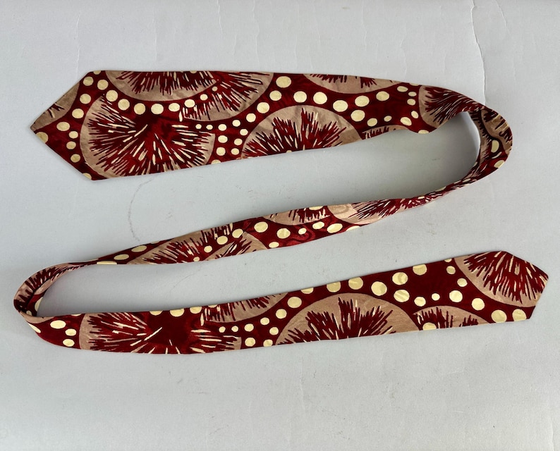 May include: A maroon necktie featuring a repeating pattern of beige half-circles, white polka dots, and maroon spiky shapes. The tie is made of a smooth fabric and is displayed on a white surface.