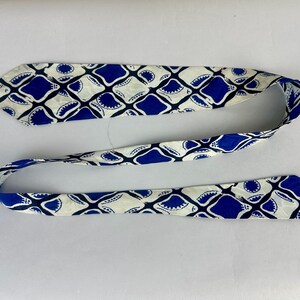 May include: A long, narrow necktie featuring a repeating geometric pattern of deep blue, rounded diamond shapes set against a cream-colored background. The design includes small, dark blue circular accents within the corners of each diamond motif.