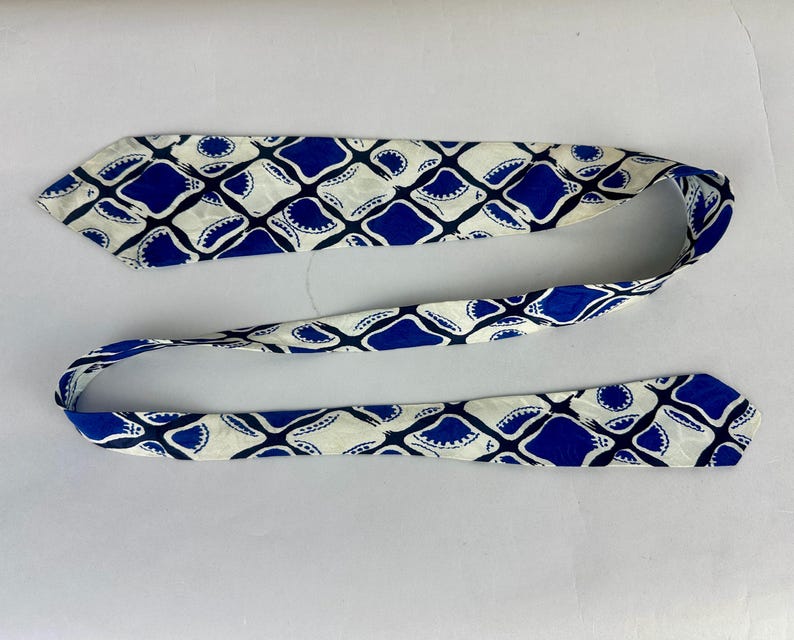 May include: A long, narrow necktie featuring a repeating geometric pattern of deep blue, rounded diamond shapes set against a cream-colored background. The design includes small, dark blue circular accents within the corners of each diamond motif.