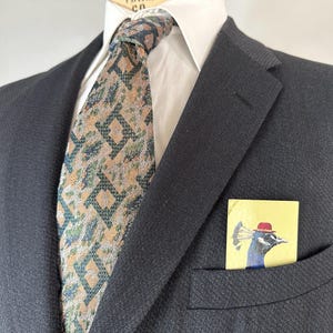 May include: A dark charcoal blazer worn over a white collared shirt with a tan and dark green geometric patterned necktie. A rectangular pocket square featuring a graphic illustration of a peacock wearing a red hat is tucked into the blazer pocket.