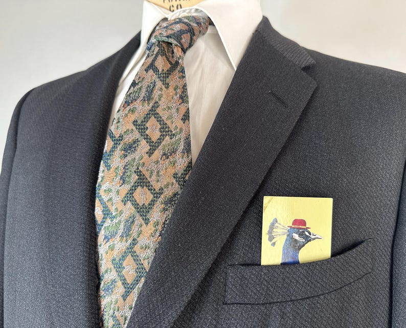 May include: A dark charcoal blazer worn over a white collared shirt with a tan and dark green geometric patterned necktie. A rectangular pocket square featuring a graphic illustration of a peacock wearing a red hat is tucked into the blazer pocket.