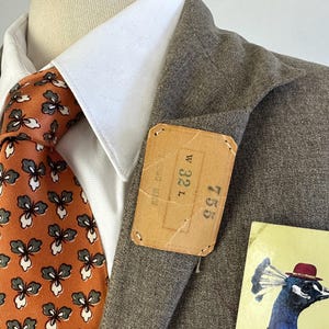 Op de afbeelding: Een bruine blazer gedragen over een wit overhemd met kraag en een oranje das met een herhalend grijs en wit bloemenpatroon. Een rechthoekig beige label is aan de revers bevestigd met de tekst W 32 L en 755. Een kaart toont een pauw met een hoed.