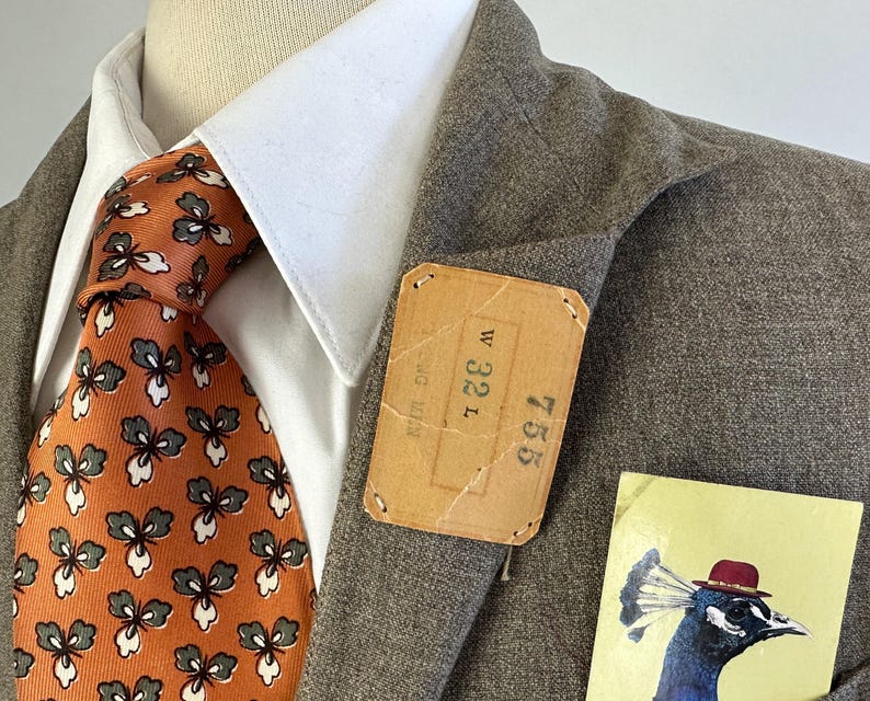 Op de afbeelding: Een bruine blazer gedragen over een wit overhemd met kraag en een oranje das met een herhalend grijs en wit bloemenpatroon. Een rechthoekig beige label is aan de revers bevestigd met de tekst W 32 L en 755. Een kaart toont een pauw met een hoed.
