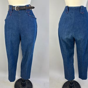 May include: High-waisted blue denim trousers featuring a dark brown leather belt with a silver-toned buckle. The pants have a structured waistband with belt loops, deep side pockets with red stitching accents, and a tapered leg silhouette.