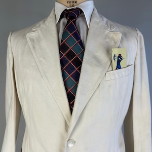 May include: An off-white blazer with a white collared shirt and a dark blue, green, and red plaid necktie. A small rectangular pocket square featuring a graphic illustration of a peacock wearing a red hat is tucked into the chest pocket.