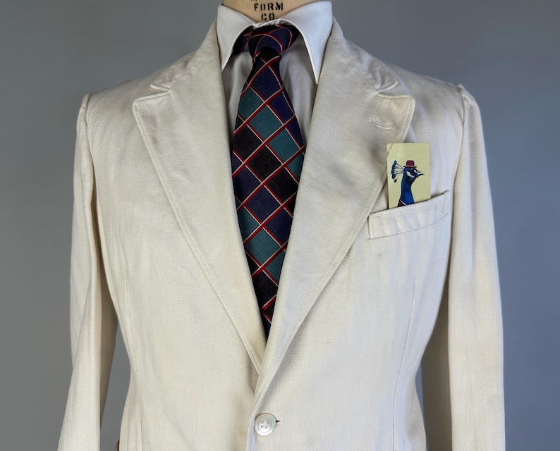 May include: An off-white blazer with a white collared shirt and a dark blue, green, and red plaid necktie. A small rectangular pocket square featuring a graphic illustration of a peacock wearing a red hat is tucked into the chest pocket.