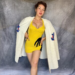 May include: A yellow sleeveless romper featuring a black hand-shaped graphic on the side, worn with a cream-colored textured cardigan that has blue and red anchor embroidery on the sleeves. The romper has a sweetheart neckline and thin straps.