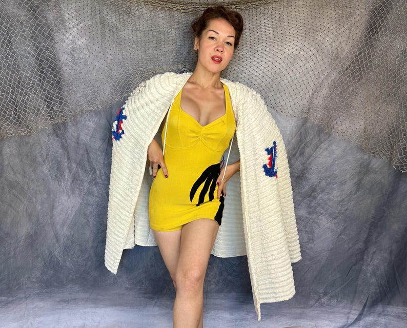 May include: A yellow sleeveless romper featuring a black hand-shaped graphic on the side, worn with a cream-colored textured cardigan that has blue and red anchor embroidery on the sleeves. The romper has a sweetheart neckline and thin straps.