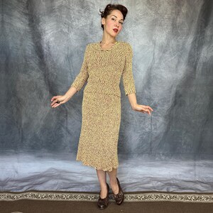 May include: A vintage two-piece dress set in a textured knit fabric. The set includes a long-sleeved top and a knee-length skirt, both in a beige color with a subtle pattern of red and brown flecks. The dress has a scalloped hem.